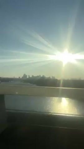 The video shows a distant cityscape over a body of water, viewed from a bridge. The sun is bright and creates lens flares, making the city skyline appear as a dark silhouette. The water reflects the sunlight. The bridge railing is in the foreground, and there is some graffiti visible on it. The scene is brightly lit and appears to be taken during the day.