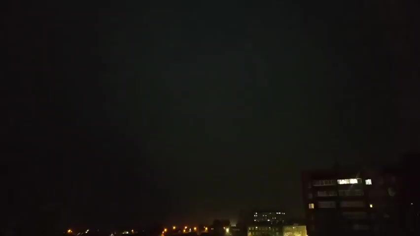 The video captures a nighttime cityscape under a dark, overcast sky. In the lower right quadrant, a multi-story brick building with illuminated windows is visible. In the distance, a line of streetlights glows, illuminating the lower portion of the frame. The sky occasionally flashes with lightning, briefly illuminating the clouds and the silhouette of buildings and trees.