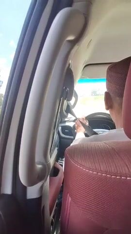 A man wearing a kufi and a white shirt is driving a car. He is wearing his seatbelt. The interior of the car features maroon and white upholstery.  The sky is visible through the windshield. There is a grab handle to the side of the passenger.  The car appears to be driving on a road lined with trees.