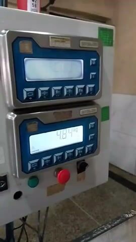 The video opens with a close-up of a control panel with two digital displays, each with a keypad. The top display shows "0.000" and the bottom shows "4.78". Below the displays are several buttons, including a red emergency stop button. The camera then pans to the right, showing a glimpse of industrial equipment and a blurred background. The next shot focuses on a stainless steel nozzle dispensing a thick, white liquid into a large cardboard box lined with plastic. The liquid creates ripples as it fills the box. The final shots continue to show the dispensing process.