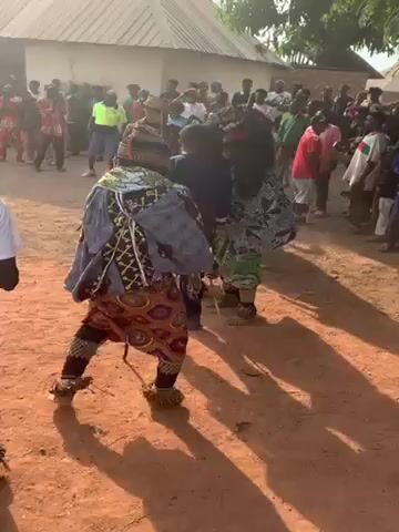 The video shows three men dancing in a circle on a dirt ground. Each dancer is wearing a colorful costume and a large mask with black hair. The dancer in the foreground is wearing a grey jacket with a geometric pattern and a brown and red patterned skirt. The other two dancers are wearing similar masks but different outfits. One is wearing a green and white patterned outfit and the other is wearing an outfit with orange and blue geometric patterns. A man in a dark suit watches them. A crowd of people are standing behind them, watching. There is a building in the background. The video appears to have been taken during the day.