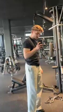 A young man stands in a gym, taking a selfie of his bicep. He is wearing a black t-shirt and light gray, baggy pants. He has brown hair styled with a side part. The gym has black walls, gray floors, and various weight machines. The man is standing near a cable machine.  A mirror is visible in the background, reflecting the gym equipment. The man flexes his right arm while looking at his phone screen.