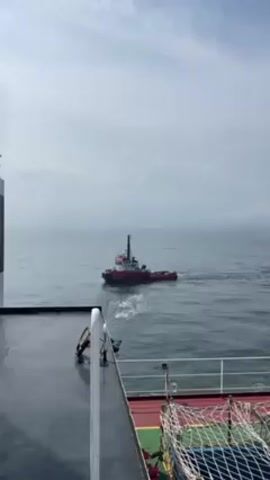 The video shows a tugboat from the deck of a larger ship. The tugboat is red and black and is moving away from the ship, creating a wake in the water. The sky is overcast.