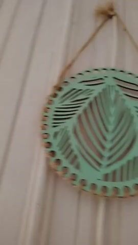 This is a close-up, slow-motion video of a wall decorated with two hanging ornaments. The wall has a textured surface with vertical lines, appearing to be wood paneling painted white. The ornaments are circular, laser-cut wooden pieces painted a light teal color. Each ornament features an intricate leaf design cut out from the center. The outer edge of each ornament is decorated with small holes, and they are bound with twine. The two ornaments are suspended by natural twine strings, creating a vertical arrangement. The top ornament is hung from a single loop of twine tied at the top. The second ornament hangs below it, attached to the bottom of the top ornament by a knot in the twine. The lighting is soft, and the camera slowly pans down the wall, revealing more of the second ornament as it moves.