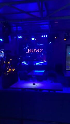 A dimly lit interior space, likely a club or lounge, is illuminated by vibrant blue lights. In the center, a large screen displays a glowing neon "NUVO" logo with abstract blue light streaks. Below the screen are several tables, one in the foreground with a hookah and a second, larger table center-screen with chairs around it. To the left, a bar area is visible with bottles on shelves. The overall ambiance is dark and moody, with pulsating lights suggesting a lively atmosphere. The video alternates between the "NUVO" logo display and a silhouette of three people standing behind a DJ setup on stage.