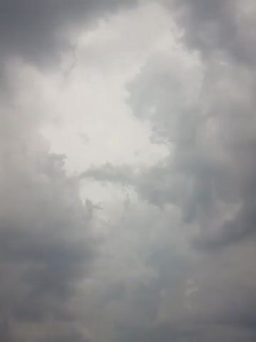 The video shows a sky filled with dark, ominous clouds. The clouds appear to be moving, indicating a potentially stormy weather condition. There is a hint of a light source somewhere above, possibly breaking through the clouds. In the lower right corner of the frame, a tall street lamp with multiple light fixtures is visible. Towards the bottom of the frame, there are also some dark shapes that could be buildings or other structures. The overall atmosphere of the video is gloomy and suggests the approach of a storm.