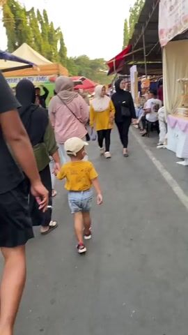 A group of people, including a young child in a yellow shirt and denim shorts, walks along a paved path lined with market stalls. The child is in the foreground, walking away from the camera. To the child's left, a person in a green shirt and striped pants is also walking. To the child's right and slightly ahead, two women in hijabs are walking, one in a pink shirt and the other in a yellow shirt. The market stalls appear to be selling various items, with tents and awnings visible. In the background, other people are browsing the stalls, and trees are visible above the awnings. The lighting suggests it is daytime, possibly late afternoon.