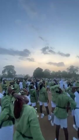 The video shows a large group of people, mostly women, in what appears to be an outdoor training or assembly area. They are all wearing matching light green shirts and white shorts, with some wearing white t-shirts underneath. Many of them have their hands on the back of their heads, as if stretching or performing a specific exercise. The sky in the background is cloudy and appears to be either early morning or late afternoon, with a soft, diffused light. Trees and some structures can be seen in the distance. The ground is a dirt or gravel surface. The overall impression is of a structured group activity taking place in an open field.