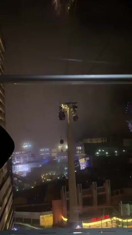 The video shows a view from inside a cable car at night.  Another cable car can be seen approaching along the cable, supported by a tall tower.  In the distance, there are lights from buildings and other structures. The sky is dark and appears to be foggy or cloudy. A person wearing a blue shirt can be seen seated in the cable car.  There are other indistinct figures in the car, as well.  The video appears to be taken from a moving cable car. The quality of the video is a bit grainy.