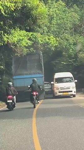 Two motorcyclists are riding behind a large blue truck on a paved road. The truck is in the right lane of a two-lane road. The motorcyclists are in the left lane. The road is surrounded by dense green foliage. The sky is not visible. The time stamp is 00:00 to 00:04.