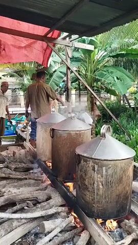 The video shows three men cooking outdoors. Three large cylindrical metal pots with conical lids are arranged on a long metal rail over a wood-burning fire. Smoke rises from the pots. One man, wearing a brown shirt and blue jeans, stands behind the pots, stirring something in the middle pot with a long utensil. Another man, wearing a beige shirt and light-colored pants, stands to the left, observing. A third man, also wearing a beige shirt, is visible behind the second man, partially obscured. The cooking area is covered by a red tarp, and lush green plants and trees are visible in the background, suggesting an outdoor or semi-outdoor setting.