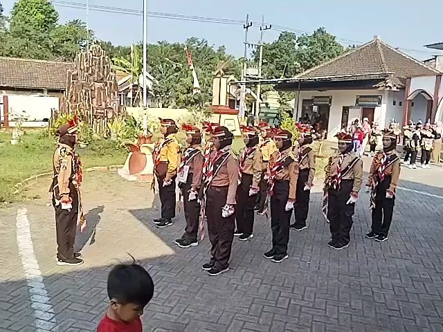 The video shows a group of young people in scout uniforms standing in formation outdoors. There are at least 15 individuals visible, all wearing brown shirts, brown neckerchiefs, and dark pants. Some have hats on. One person, seemingly an instructor or leader, stands facing the group, also in a uniform. The group appears to be in a courtyard or open area with a paved surface made of interlocking bricks. In the background, there are buildings, trees, and a flagpole. The sky is clear and the scene is well-lit, suggesting it is daytime. The individuals are standing at attention, with some appearing to be responding to commands from the leader. The overall atmosphere seems formal and organized, likely related to a scout activity or ceremony.