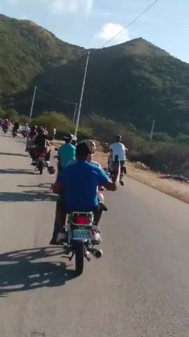 The video shows a group of people riding motorcycles on a paved road. The camera is positioned behind one of the motorcycles, offering a rear view of the riders ahead. Most of the motorcyclists are wearing casual clothing like t-shirts, and some have helmets. The motorcycles appear to be small, similar in style, and are moving at a moderate speed.

The road is situated in a rural area, with green, mountainous terrain visible in the background. There are also tall, thin lampposts lining the roadside. The sky is light blue with scattered clouds, suggesting a sunny day. The wind is audible in the video. Some of the riders are waving or gesturing with their hands as they ride.