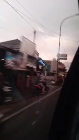 A car is moving slowly down a street lined with shops and buildings, the camera positioned inside, looking out the driver's side window. The day is overcast, and the road appears slightly wet, perhaps from recent rain. A silver minivan is partially visible on the left side of the road, alongside a building with an awning.  Further down the street, there are several other shops and buildings.  A food cart is on the right side of the street, and there are people on the sidewalk near the shops.  Electrical wires stretch overhead, and there is a sign for "Indomaret" visible in the distance.  A person on a motorcycle is moving along the road in the opposite direction of the car.  The overall impression is of a quiet, typical street scene in a small town.