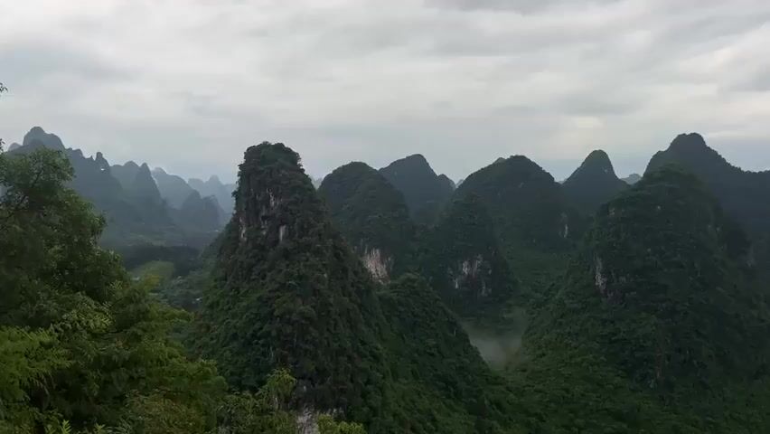 The video displays a panoramic view of lush green mountains, possibly of a karst landscape, under a cloudy sky. The mountains appear to be covered in dense vegetation, and their unique, conical shapes create a distinctive skyline. The visibility extends far into the distance, with additional mountains gradually fading into the horizon. In the distance, the presence of a river and a small settlement can be observed, adding a sense of scale to the landscape. No people are directly visible in the frame, though laughter or talking can be heard.