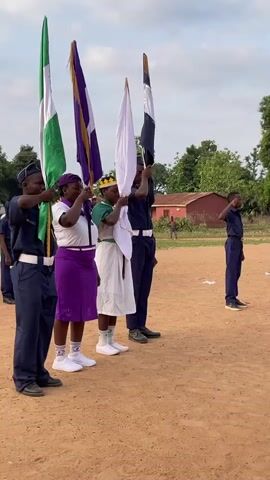 A group of people in uniforms are standing on a dirt field, holding flags. The uniforms are navy blue with white belts and caps. One person wears a white dress with a green sash, a crown, and white socks with white sneakers. One flag is the Nigerian flag. The second flag is purple and white. The third flag is white. The fourth flag is black and white. The people are standing in formation and holding the flags up. Another person in a navy uniform stands to the right and salutes. In the background, there is a building and some trees. A child walks around in the background.