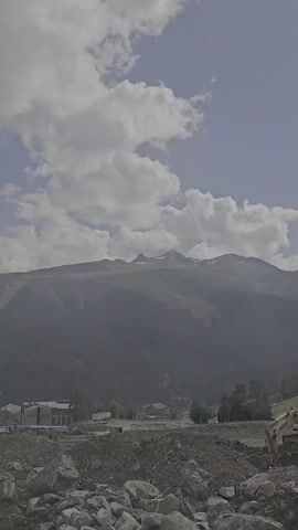 The video shows a mountain landscape on a cloudy day. In the foreground, there is a pile of rocks and dirt, and some construction equipment can be seen on the right side. In the middle ground, there are several buildings, some trees, and a forested slope. In the background, there are large mountains, some with snow-capped peaks. The sky is blue with white clouds.