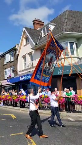This is a first-person perspective video of a parade marching down a street lined with shops and buildings. The sky is blue with some clouds. People are gathered on the sidewalk watching the parade. The parade participants are dressed in matching uniforms, predominantly white shirts with blue pants and sashes. They are carrying flags and banners. Two individuals in the foreground are holding a large orange and blue banner with imagery on it. The street has yellow lane markings. The shops visible include "BoyleSports." The overall atmosphere appears to be festive.