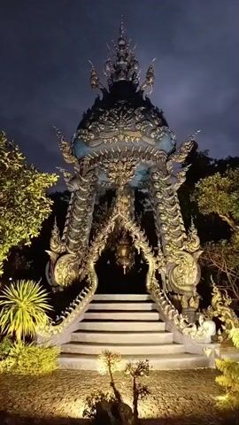 The video shows an ornate, illuminated structure at night. The structure is tall and intricate, with a tiered, conical roof adorned with golden decorative elements. It is supported by a series of curving, dragon-like sculptures that wrap around the central opening. A set of stairs leads up to the entrance of the structure, which is dimly lit from within. The ground in front of the structure is covered in gravel and illuminated by spotlights, highlighting small bonsai trees and rocks. Lush green foliage surrounds the structure, and the night sky is visible above. The overall impression is one of a beautifully lit, ancient, and possibly religious monument.