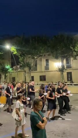 A group of people dressed in black are performing outdoors at night. They are holding and playing hand drums and tambourines, suggesting they are a musical ensemble. The background shows a building with lights on and trees lining the street. Spectators are standing on the side, observing the performance. The overall atmosphere appears to be a lively outdoor event.