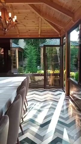 This video showcases an outdoor seating area with a wooden roof and glass walls, overlooking a lush garden. The floor features a striking chevron pattern in shades of white, grey, and teal. Several upholstered chairs are visible along one wall, hinting at a dining or lounge setup.

The garden beyond is a vibrant green, filled with various trees and manicured bushes. A small, charming gazebo with a green roof is nestled amongst the foliage. In the distance, two individuals are seated on a wooden bench, appearing relaxed and engaged in conversation. String lights are visible throughout the garden, suggesting the possibility of evening use.

The scene appears to be set during daylight hours, with natural light illuminating the area. The overall atmosphere is serene and inviting, suggesting a peaceful retreat or an extension of an indoor living space. The glass walls allow for an unimpeded view of the natural beauty outside, blurring the lines between indoor comfort and the outdoors. The polished floor reflects the light and the greenery, adding to the sense of spaciousness and elegance.

While no explicit text is visible, the setting itself conveys a sense of refined leisure and an appreciation for nature. The location context provided, Buzovna, Azerbaijan, suggests a potentially temperate climate suitable for enjoying such an outdoor space.
