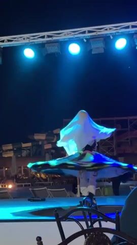 A dancer performs a whirling dervish dance on a blue-lit stage at night. The dancer wears a white head covering, a patterned vest, light-colored pants, and a large, colorful skirt that spins out during the dance. Behind the stage are structures and beach chairs, suggesting an outdoor venue. The lighting rig above the stage includes multiple spotlights. A few audience members sit at tables in the foreground.
