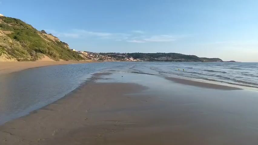 The video shows a coastal landscape at what appears to be either sunrise or sunset. The scene features a sandy beach with a body of water in the foreground, possibly the mouth of a river or stream meeting the sea. The beach is mostly flat and wet, reflecting the sky and sun. 

In the distance, a town or settlement is visible along the coastline. A headland extends into the sea on the right side of the frame. The sun is positioned in the sky, creating a bright glare and reflecting on the water. There are a few people visible in the distance in the water. 

The sky is blue with some clouds. The general lighting suggests a calm and peaceful environment.