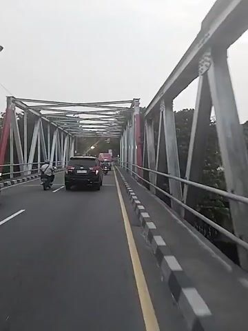 The video shows a view from a vehicle moving across a metal truss bridge. Several vehicles including a black SUV and motorcycles are on the bridge. The sky is overcast and trees are visible on either side of the bridge. The bridge structure is painted silver and red.  The road is divided by a yellow line, with a black and white checkered curb along the side.