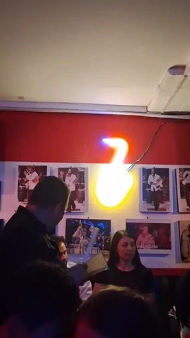 The video shows the interior of a bar. There are several people, mostly in dark clothing, standing or sitting at tables. A man in a black shirt appears to be taking an order from a woman. The bar features red walls with framed photos of musicians and a neon light shaped like a musical note. Above the patrons, a television screen shows bright, colorful graphics. A bar area with a "salsa clásica" sign and a refrigerated drinks display is visible, along with stained-glass decorations. The lighting is dim and the camera movements are somewhat shaky, giving an impression of a lively, intimate setting.