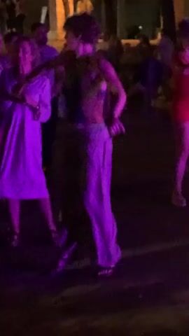 The video shows a nighttime outdoor scene with several people dancing under purple lighting. In the foreground, two women are dancing together. The woman on the left is wearing a long white dress and the woman on the right is wearing a dark top with a patterned design and loose-fitting light-colored pants. They are holding hands and appear to be engaged in a lively dance. Other people are visible in the background, also dancing and socializing. The environment appears to be some sort of public space, possibly a plaza or park, with structures and trees dimly lit in the background. The overall mood is festive and energetic.