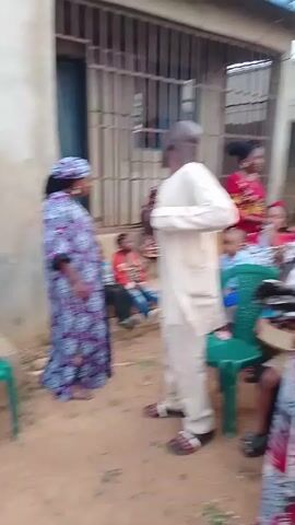 A group of people gather in an outdoor space. They are mostly dressed in colorful African-style clothing and some are carrying handbags or trays. A few people are sitting on plastic chairs. The area is partially enclosed by buildings and walls. There's a papaya tree in the background. The people seem to be talking amongst themselves.