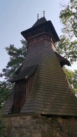 The video shows a close-up, low-angle shot of a wooden bell tower situated outdoors. The tower has a steep, shingled roof and a stone base. Trees with green foliage are visible in the background against a bright, clear sky. The camera angle is fixed, and there is no discernible movement or audio in the brief clip. The structure appears to be weathered and rustic, suggesting it is an older building, possibly in a rural or historical setting. No people are visible in the frame.