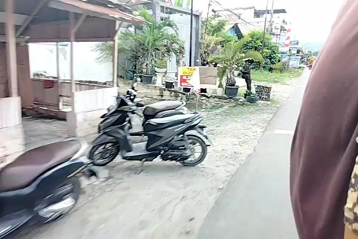 The video shows a first-person perspective of someone riding on a vehicle, likely a motorcycle or a bicycle, through a street. The street appears to be in a rural or semi-urban area, with small buildings and vegetation lining the sides. There are other people and vehicles on the street, including pedestrians and motorcycles. The overall atmosphere is casual and everyday.