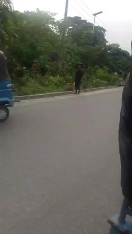 The video shows a view from a moving vehicle on a paved road with trees and vegetation on the side. A man in a dark outfit is walking along the road at the beginning of the video. Then, a black sedan passes the camera, followed by a dark blue SUV. The weather appears overcast.