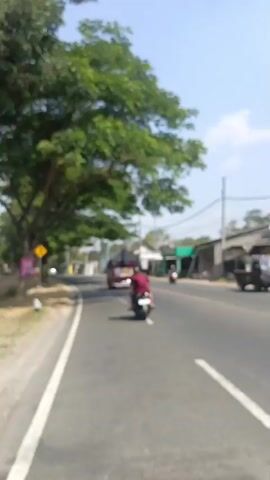 The video shows a blurred view of a road. There are trees with green leaves on the left side of the road, casting shadows on the asphalt. A yellow diamond-shaped sign is visible on the left side of the road. A motorcycle is seen riding on the road in the distance, followed by a truck. There are small buildings and shops along the right side of the road. The sky is clear and bright. The video appears to be taken from a moving vehicle, possibly another motorcycle, given the perspective.
