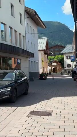 This video captures a street scene in Zell am See, Austria, on a sunny day. The camera pans slowly to the right, revealing a charming European street lined with multi-story buildings featuring traditional alpine architecture. Many of the buildings have pitched roofs, some with exposed wooden beams. One building on the left has a modern facade with large windows, reflecting the blue sky and green mountains in the distance.

The street is paved with brickwork, and shadows from the buildings and a pole stretch across it, indicating it is likely late morning or early afternoon. A dark-colored car is parked on the left, and a bicycle is leaning against a barrier on the right. In the background, a covered outdoor seating area with a sign that reads "Sonnenbank" is visible. Further up the street, a person on a bicycle is seen riding away from the camera.

Mountsaint scenery forms a picturesque backdrop to the town, with lush green slopes and a clear blue sky dotted with a few white clouds. The overall atmosphere is peaceful and serene, suggesting a typical day in this tourist-friendly Austrian town. Visible text includes "P" signs indicating parking and street signs that appear to read "Seegasse" and "9".