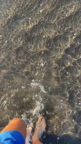 A low-angle, point-of-view shot shows a person wading through shallow water. The person is wearing bright blue shorts, and their bare foot enters the water, causing a splash. The water is relatively clear but has a murky bottom visible. Sunlight reflects off the water's surface, creating a shimmering effect. The overall scene has a natural, possibly coastal or beach-like setting.
