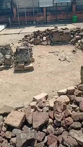 The video pans across an outdoor area with archaeological ruins. In the foreground, piles of broken bricks and stones are scattered on the dirt ground, some forming low walls. In the center, two stone structures, appearing to be foundations or altars, are visible, surrounded by more brick fragments. Behind these ruins, a small, white building with a dark roof stands. A dirt path leads to the building. Two motorcycles are parked near the building, one red and white, the other black. Several people are sitting on a bench in front of the building, under a shaded overhang. The background shows a brick wall and fence, and some greenery. The overall impression is a historical site undergoing preservation or excavation.
