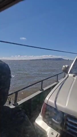 A ferry is crossing a body of water on a sunny day. The camera is positioned inside a vehicle, looking out the window at the water and the sky. A silver SUV is parked next to the vehicle. The water is choppy, and there are some whitecaps. In the distance, there is a landmass with buildings on it. The sky is a clear blue with a few scattered clouds. The ferry is moving, as evidenced by the wake behind it. The scene is peaceful and serene.