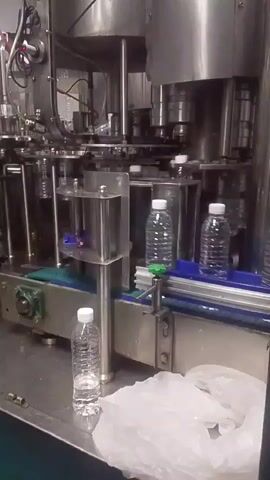 A bottling machine is shown in operation, filling and capping plastic water bottles. Bottles move along a conveyor belt toward a capping mechanism. Several bottles are visible on the belt, some already filled and capped, others in the process of being capped. The machine appears to be made of stainless steel and is likely in a factory or production environment. The video captures the automated process of bottling water.