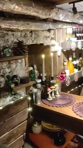 The video pans across a home bar area. The walls appear to be made of rough stone or concrete. There are shelves stocked with bottles of alcohol, some of which are illuminated by fairy lights. On the counter, there are decorative items such as mushrooms, a pink piggy bank, and a carved bust of a creature with pointed ears. A wind chime made of metallic discs hangs in the center of the frame. In the background, a record player is set up, with a record spinning. The overall atmosphere suggests a cozy, perhaps slightly quirky, personal space. There are no people visible in the video.