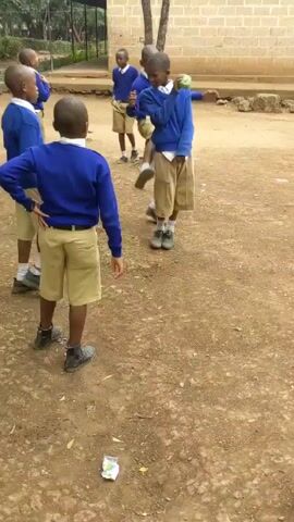 The video shows several young boys in blue sweaters and khaki shorts playing in a dirt area with a green ball. A few of the boys are actively chasing each other and throwing or passing the ball, while others are watching or standing nearby. There's a small brick building and trees in the background. A piece of litter sits on the ground in the lower-left corner.