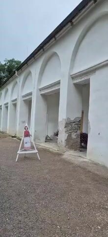 The video begins with a shot of an arched arcade with white walls. A stand with a cutout figure of a person wearing a red dress and white apron is visible in the foreground. The camera pans to the right, revealing the continuation of the arcade and a patch of stone wall with crumbling plaster. A wooden object and some dark foliage are visible in an alcove. The camera continues to pan, showing a long, yellow-ochre colored wall with arched openings and a green lawn. A tall evergreen tree stands in front of the wall. The camera then shifts to show a light yellow building with a brown roof. In front of the building, there is a ramp, a stand with a cutout figure similar to the first one, and an informational display box. The scene then moves to show a gravel path leading away from the building, with more trees and greenery on either side. A white car is seen driving down the path. Another stand with a cutout figure and a flower basket is visible near the building. The video concludes with a shot of the path and the surrounding greenery.