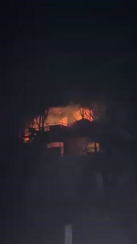 The video depicts a building engulfed in flames at night. The fire is visible through the building's structure and surrounded by smoke. Bare trees are silhouetted in the foreground, partially obscuring the view of the burning building. The sky is completely dark, and the overall environment suggests a rural or suburban setting. The footage is dark, emphasizing the intense orange glow of the fire against the night. A faint white light source illuminates the bottom right corner of the frame.