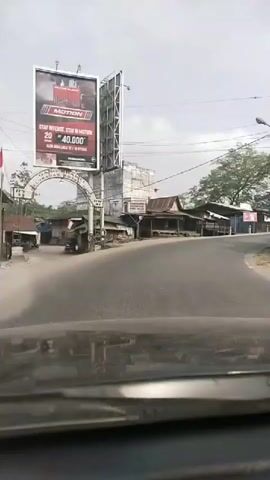 The video shows a road scene from the perspective of a vehicle's dashboard. On the left, a large billboard displays the word "MOTION" and text in Indonesian, possibly an advertisement. An arched gateway with the words "JL. SUDIRMAN NO. 103" is visible. Along the right side of the road, a series of small, simple structures with corrugated metal roofs appear to be shops or stalls. A few motorcycles are seen on the road, and trees are present in the background. The sky is overcast.