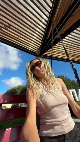 A young woman with blonde curly hair and sunglasses is sitting on a red bench. She is wearing a light-colored tank top and black denim shorts. The woman is holding a camera and appears to be taking a selfie. Behind her, there is a large sign with white letters that spell out "ПАВЛОВСК." The scene is outdoors, with trees and a blue sky visible in the background. The woman is smiling and looking at the camera.
