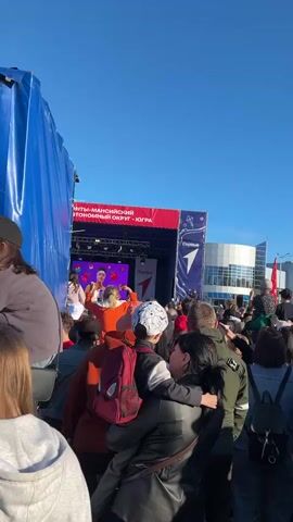The scene depicts a crowd of people gathered in front of a stage on a sunny day. The stage is decorated with red and blue banners, and a performer is visible on stage, lit by stage lights and shown on a screen. The crowd is diverse, with individuals of different ages and ethnicities. People are wearing casual clothing and some are holding up their phones, presumably to take pictures or videos. A blue tarp is visible on the left side of the frame. In the foreground, a woman is holding a child. In the background, a large, modern building is visible. The overall atmosphere appears to be festive and excited.