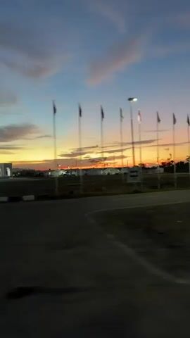 The video captures a scenic view of a sunset in Meureudu, Indonesia. The sky is ablaze with vibrant hues of orange, yellow, and pink, transitioning into a deep blue as dusk settles. Several clouds are scattered across the sky, adding texture and depth to the atmospheric spectacle.

In the foreground, a road is visible, suggesting that the footage was likely filmed from a moving vehicle. Along the roadside, a row of flagpoles stands tall, each adorned with a flag, though the details of the flags are obscured by the fading light and motion. Beyond the flags, a low-rise building complex is visible, possibly an industrial area or residential development, with some lights starting to twinkle. A single streetlamp illuminates the scene from a flagpole in the mid-ground. A large tree with blurred branches is also present, indicating the passage of time or movement.

The overall mood is serene and contemplative, highlighted by the beauty of the natural sunset. The time of day is clearly twilight, characterized by the fading sunlight and the emerging darkness. The location cues, such as the distinctive tropical foliage and the architecture of the buildings, along with the explicit mention of Meureudu, Indonesia, firmly establish the geographical context. No legible text is visible in the media.
