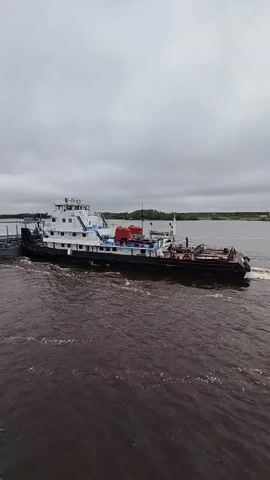 The video shows a tugboat pulling a barge on a wide river under an overcast sky. The tugboat is black and white with red details, and the barge is long and flat. The river is choppy and brown.  The camera is positioned slightly above the water level, possibly on another boat.  Trees line the shore on either side of the river.  A green handrail of the vessel from which the video is being recorded is seen in the bottom left.
