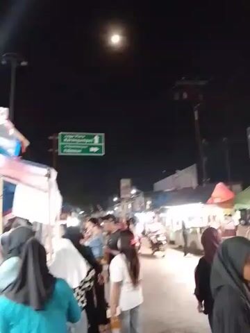 The video shows a crowded street market at night. People walk around, many wearing hijabs. There are food stalls with colorful awnings and bright lights. Motorcycles weave through the crowds. The sky is dark with a bright full moon. Streetlights illuminate the scene. A green road sign points in one direction.
